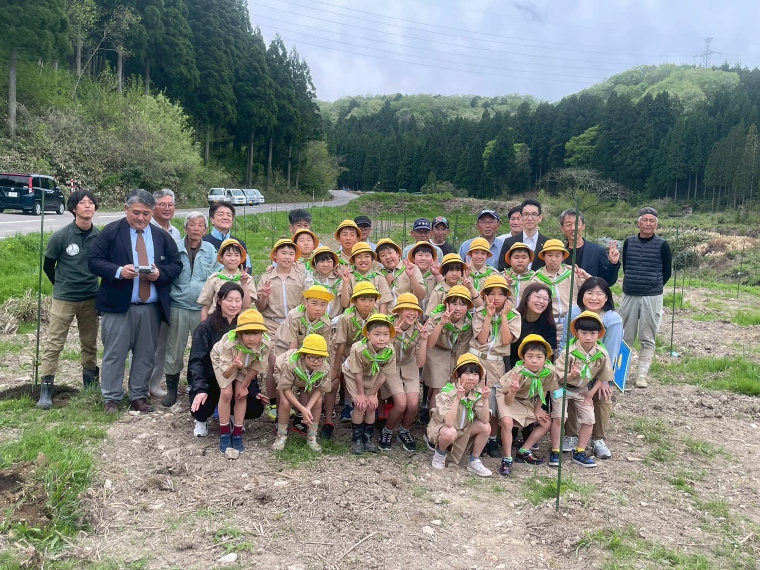 植樹後に圃場で記念撮影する那谷小学校の全学年児童と関係者。黄色い帽子と緑のスカーフの制服姿の子どもたちが笑顔でポーズをとっている