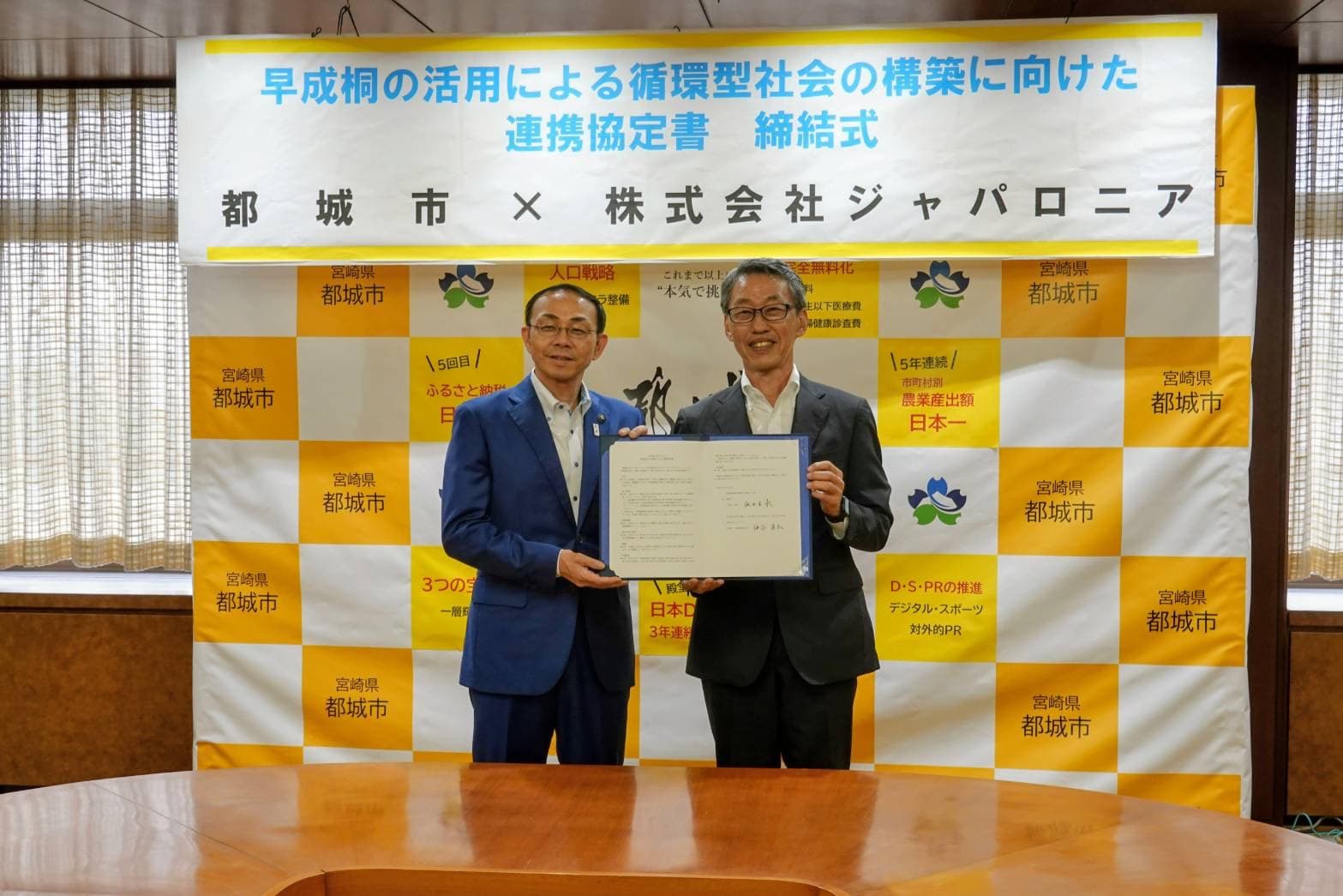 都城市とジャパロニアの連携協定締結式。バナーの前で協定書を手に持つ両者の代表者